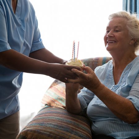 Happy senior woman looking at cup cake with candle held by nurse in nursing home